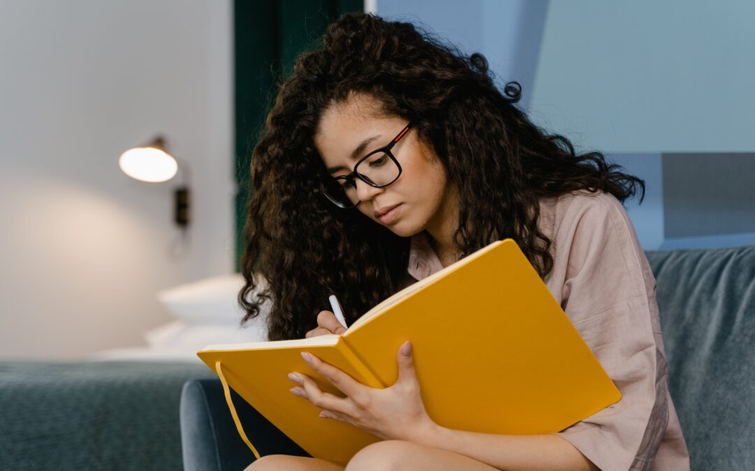 Woman with black hair and glasses wearing glasses, holding an open journal and a pen